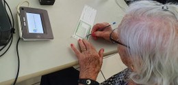 Fotografia de uma senhora de cabelos brancos assinando um título de eleitor sobre a mesa. À esqu...